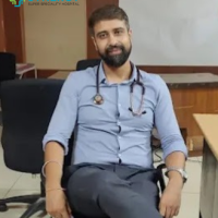 Expert Doctor at Synergy Hospital Doctor sitting in an office chair, wearing a light blue shirt and stethoscope, representing Synergy Hospital's expert medical team."